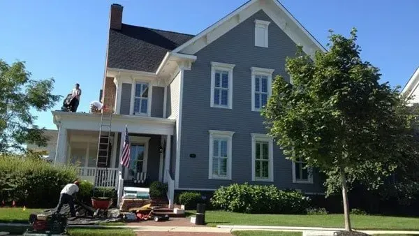 Workers on roof of two-story house, painting. Blue siding, white trim. American flag. Sunny day.