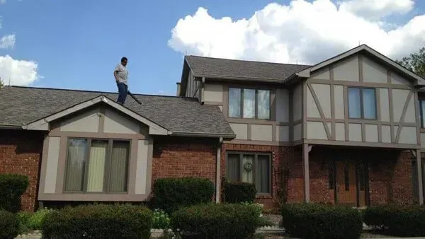 Person blowing leaves off a roof of a two-story brick house with brown trim and blue sky.