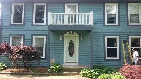 Two-story blue house with white trim. A person on a ladder is painting. Small balcony above front door.