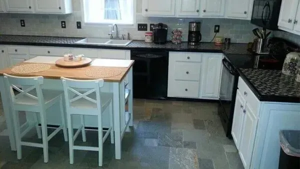 Kitchen with white cabinets, black countertops, island with two white chairs, and gray flooring.
