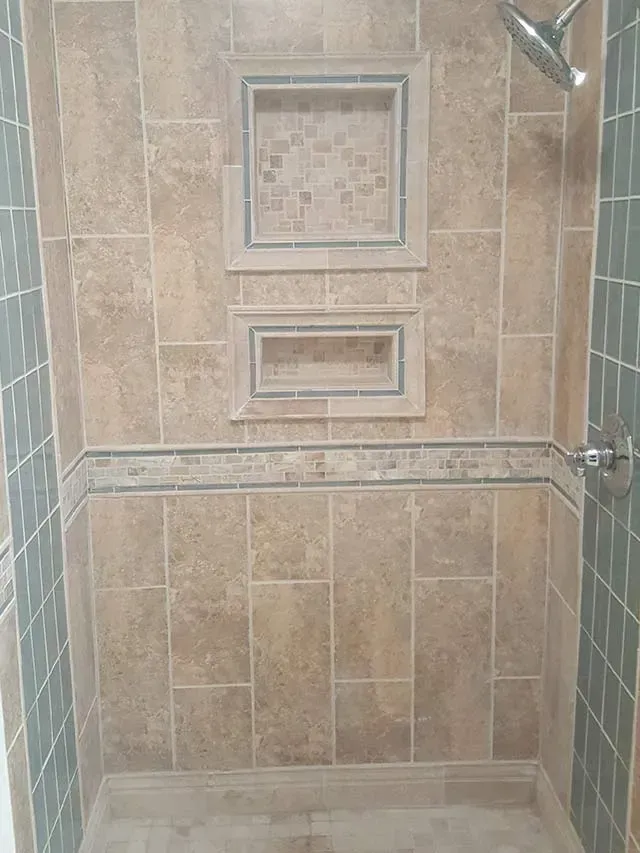 Shower tiled in beige and turquoise, featuring two built-in storage niches with mosaic tile accents and a shower head.