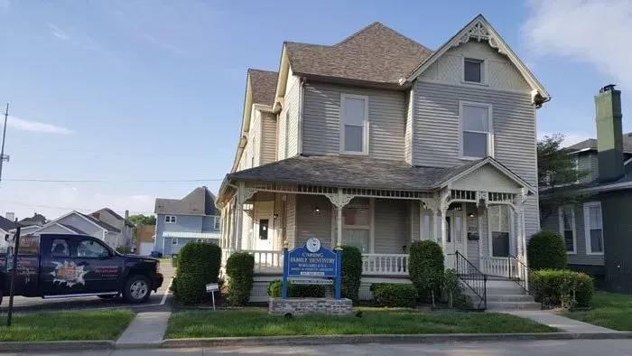 Two-story gray house with porch; sign in front, blue truck parked on the left.