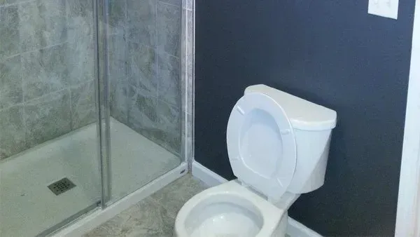 Bathroom with a toilet, shower with glass doors, and gray tiled walls. The toilet seat is up and walls are dark blue.
