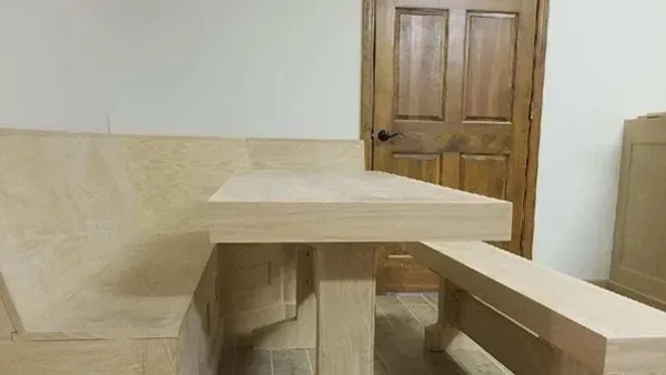 Light wood dining table and bench in front of built-in stone booth seating. Wooden door in background.