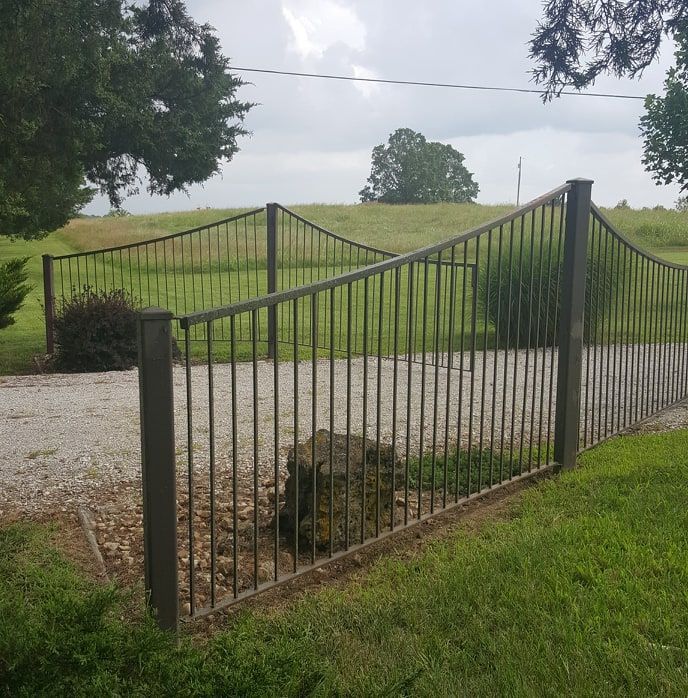 A metal fence surrounds a gravel driveway leading to a grassy field