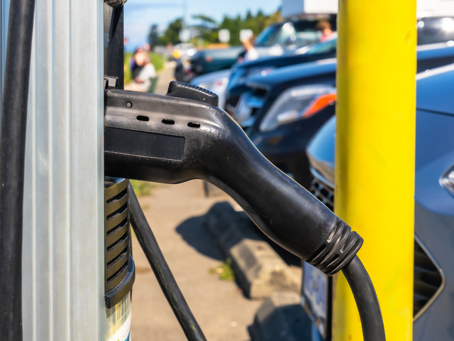Black charging cable plugged into an electric vehicle charging station, parked cars in background.