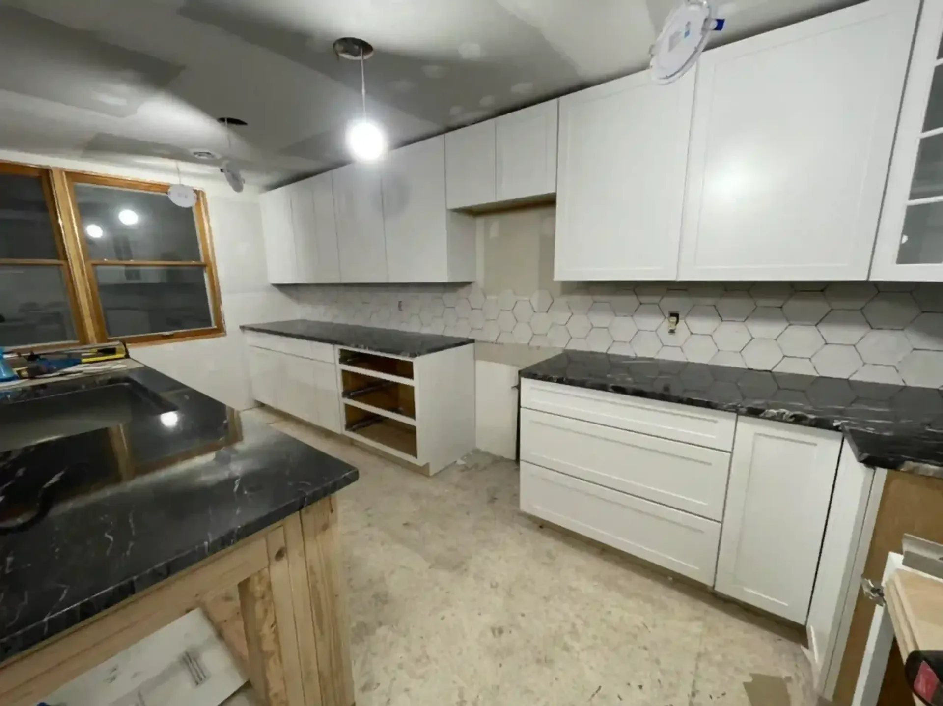 Kitchen under renovation; white cabinets, black countertops, hexagon tile backsplash, unfinished floor.