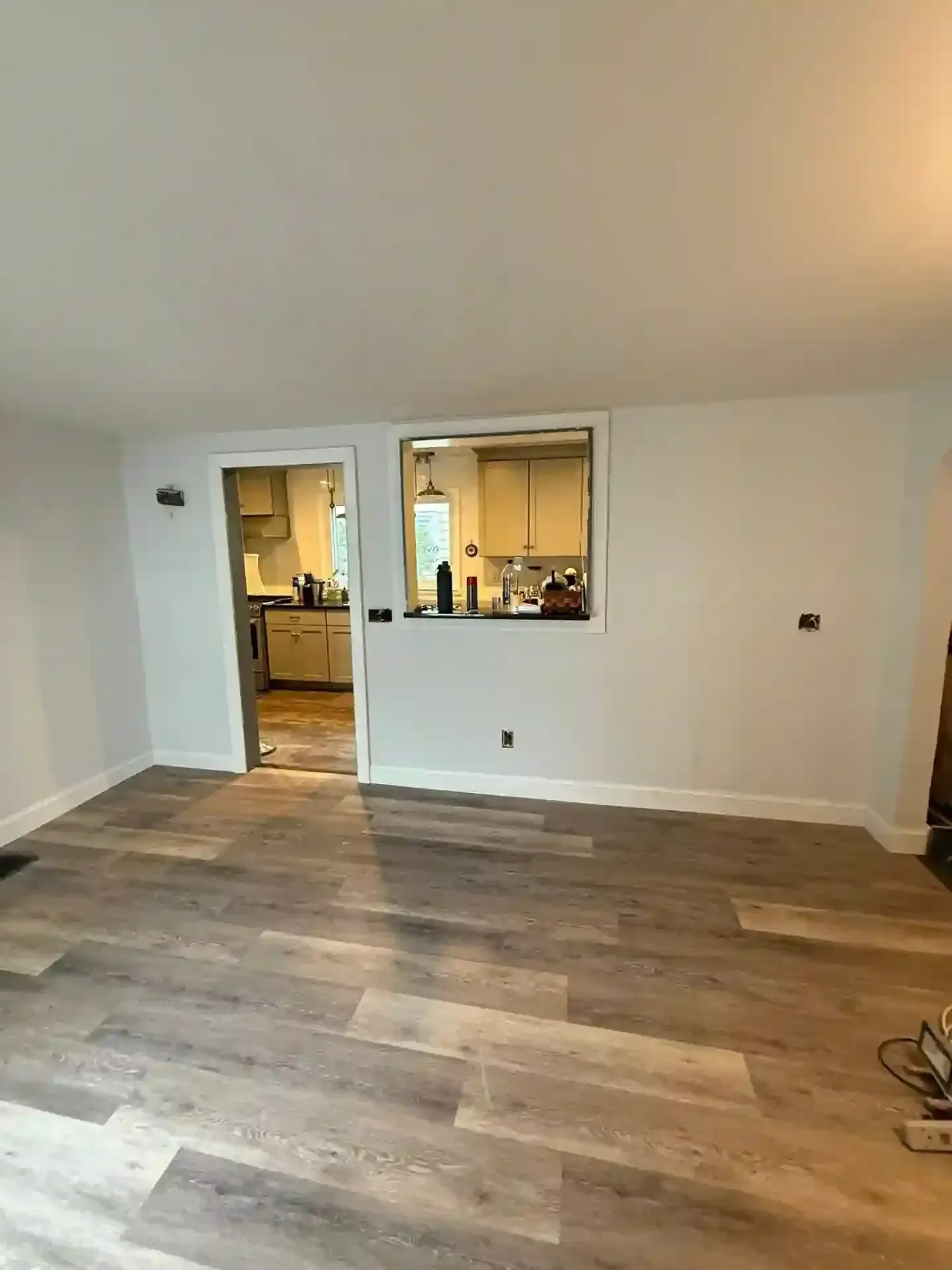 Empty living room with wood-look flooring, door to a kitchen, and a pass-through window. White walls.