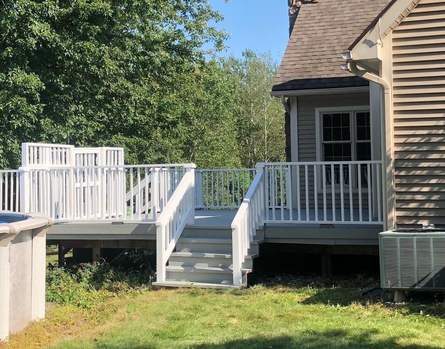 White deck with stairs, built against a house with a gray roof and siding. Green grass and trees in the background.