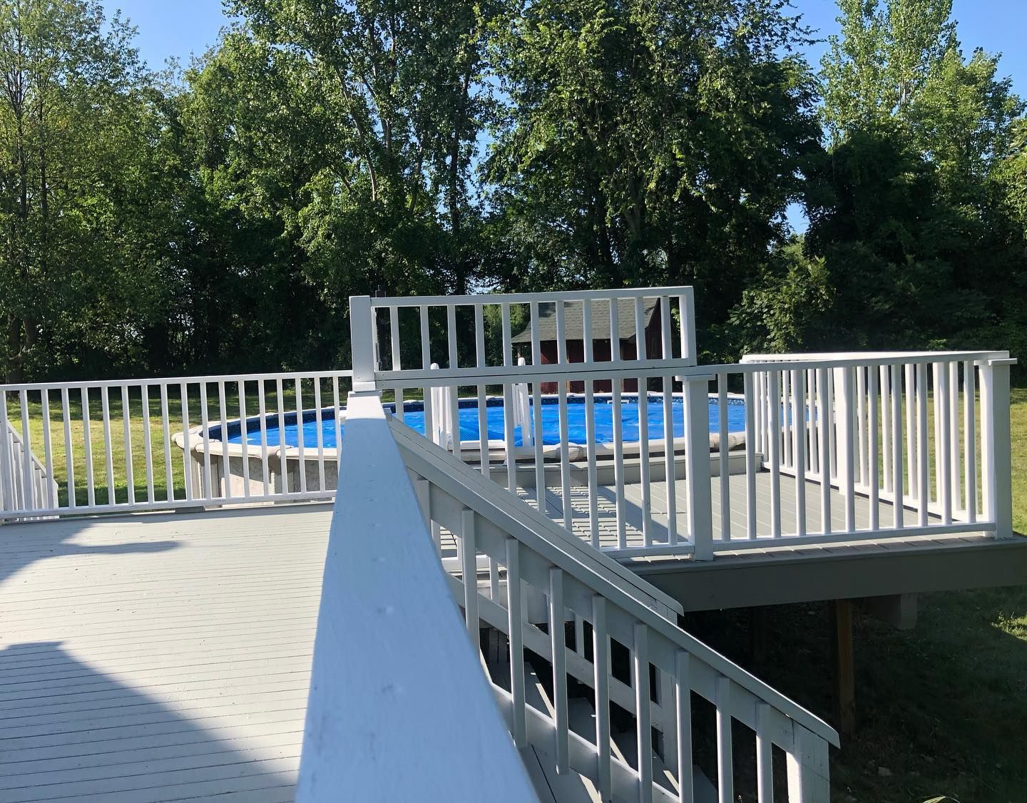 White deck with a white-railed fence surrounds an above-ground pool with a blue cover, surrounded by green trees.