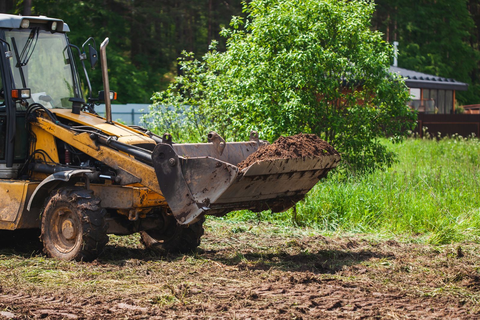 Yellow tractor with a full bucket of dirt, on a brown, cleared field near greenery.