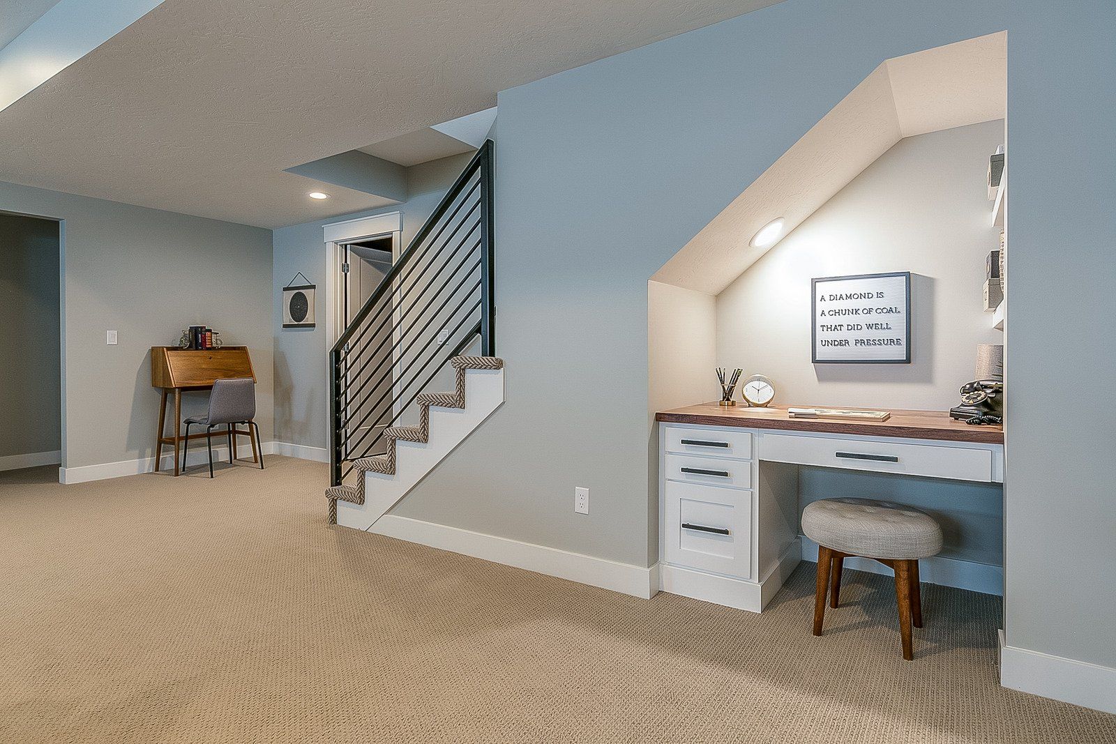 Basement interior with stairs, a desk under the stairs, and a rug. Gray walls and carpet.