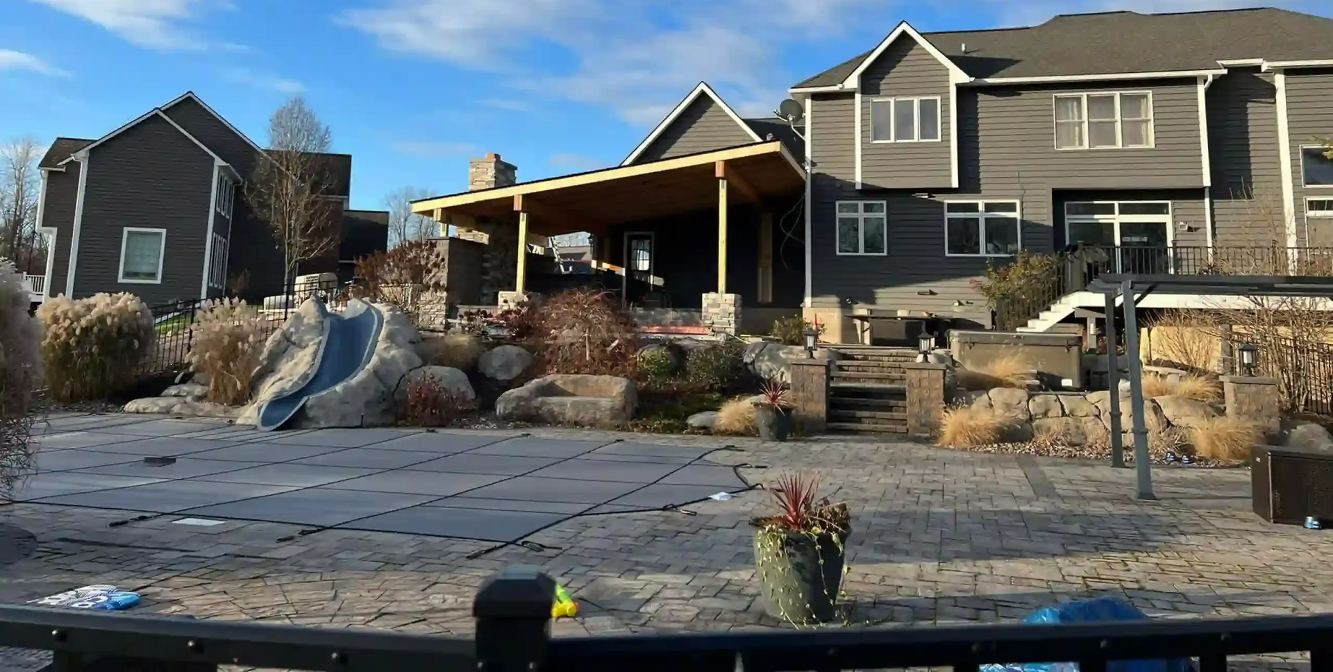 A gray deck with white railing and stairs next to a house with a green bush.