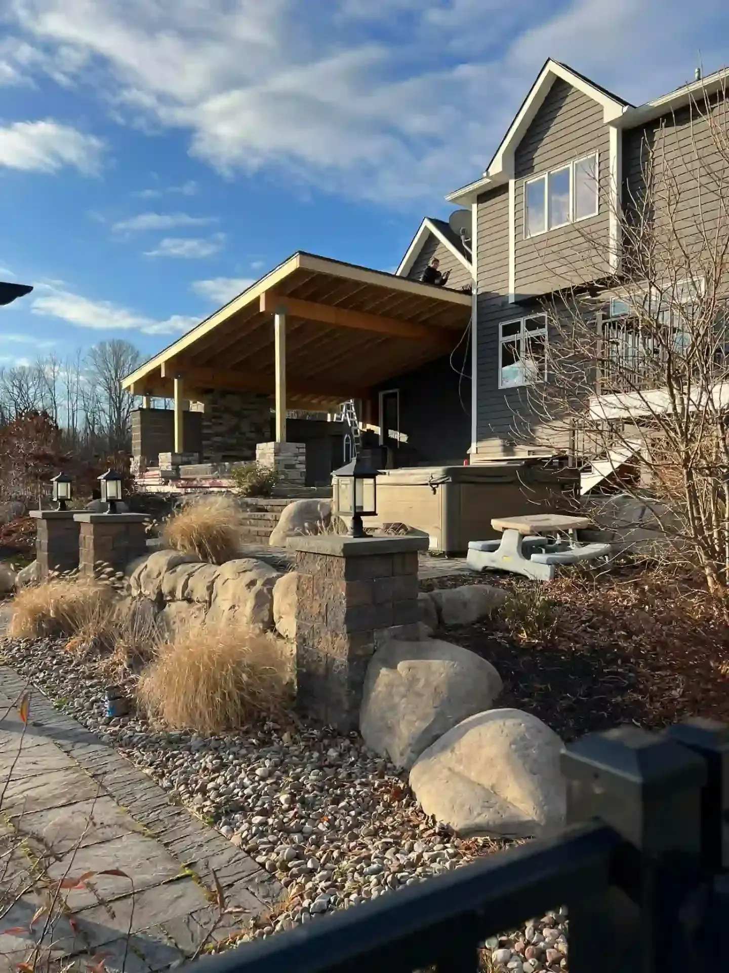 Patio with wooden canopy, rock wall, large rocks, and a two-story house under a blue sky.