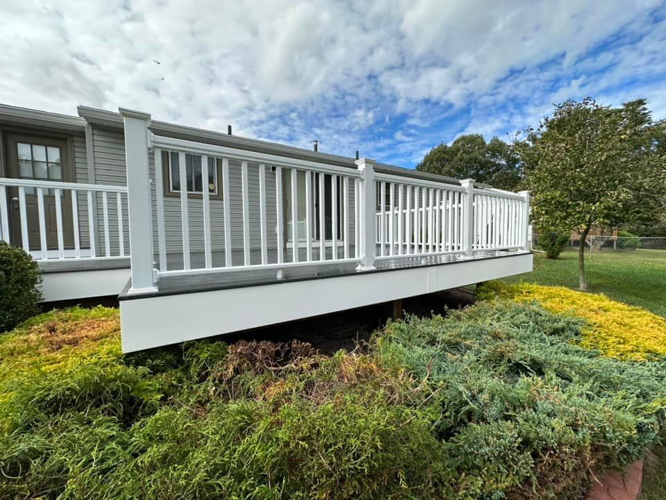 White deck with railing attached to a light gray house. Green and yellow bushes in front.