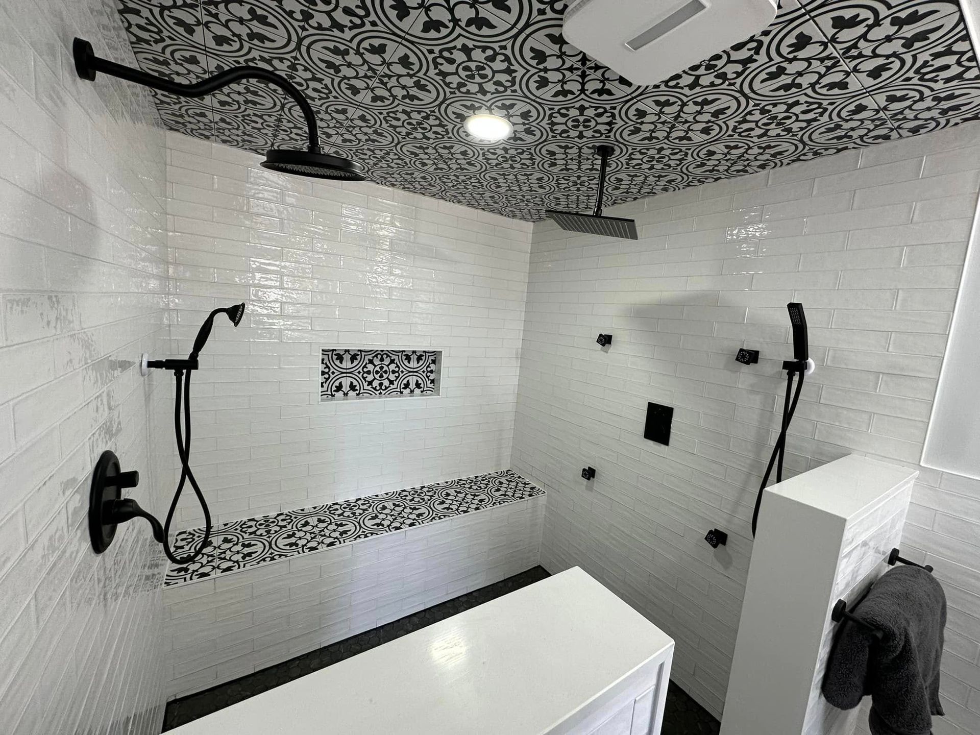 Black and white tiled shower with overhead and handheld shower heads, bench seat, patterned ceiling.