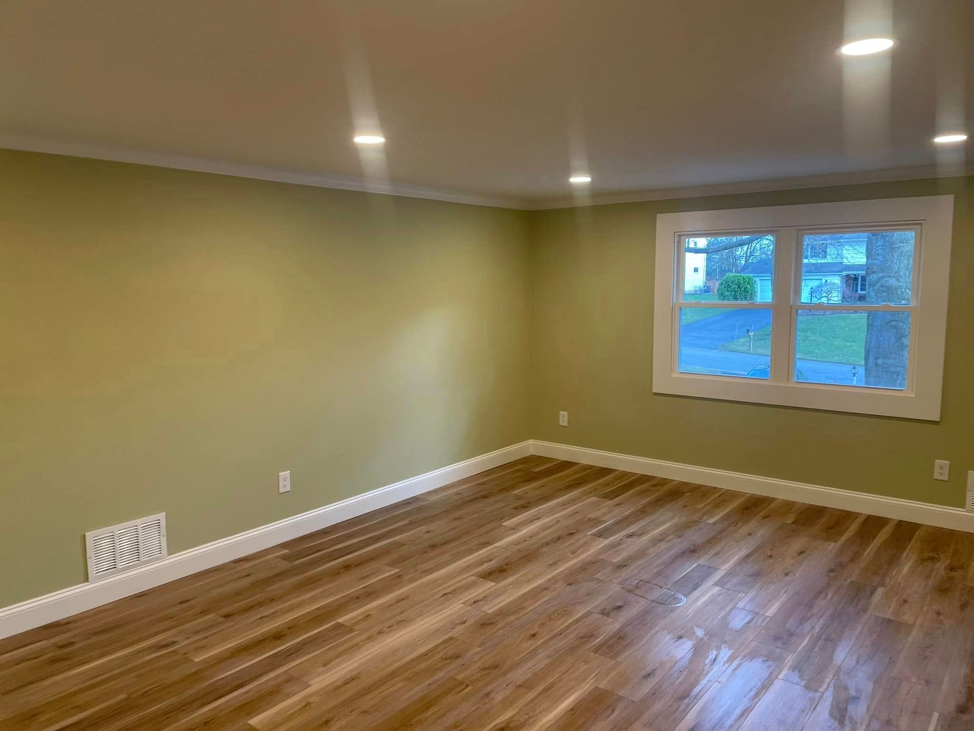 Empty room with wooden floor, olive green walls, white trim, and a window.