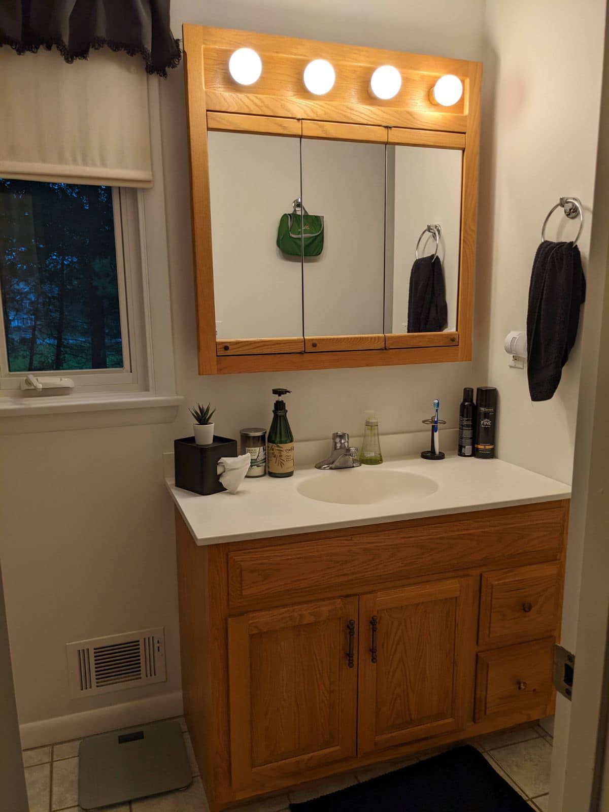 Bathroom with wooden vanity, mirror, and four-light fixture. A window with a shade and a towel holder are also visible.