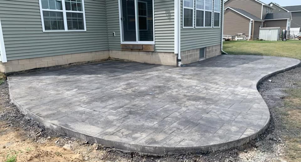 Gray paver patio next to a house with a rounded edge and unpaved surrounding dirt.