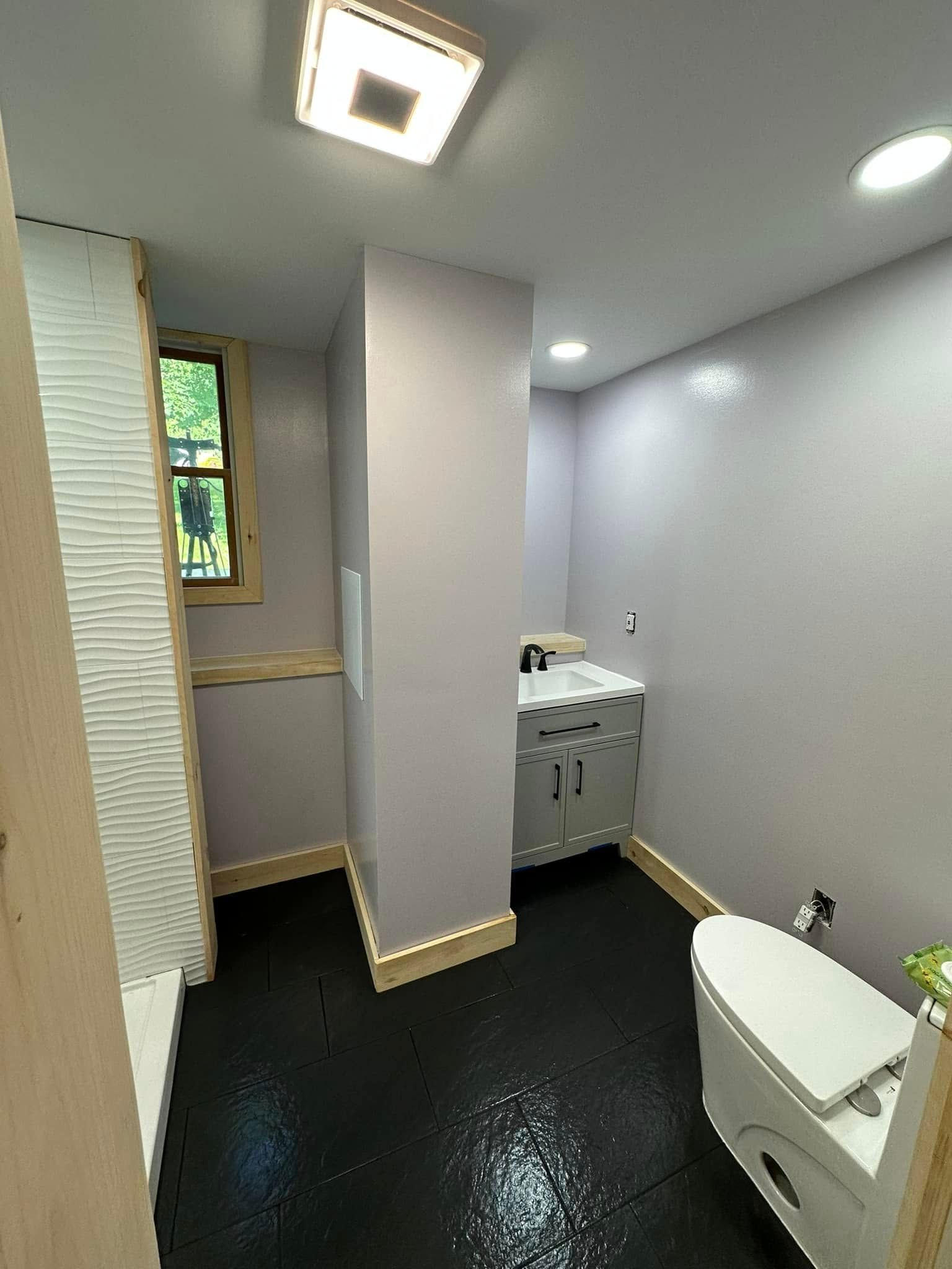 Small bathroom with black floor, toilet, vanity, and window. Walls are purple-gray. Bright overhead lighting.