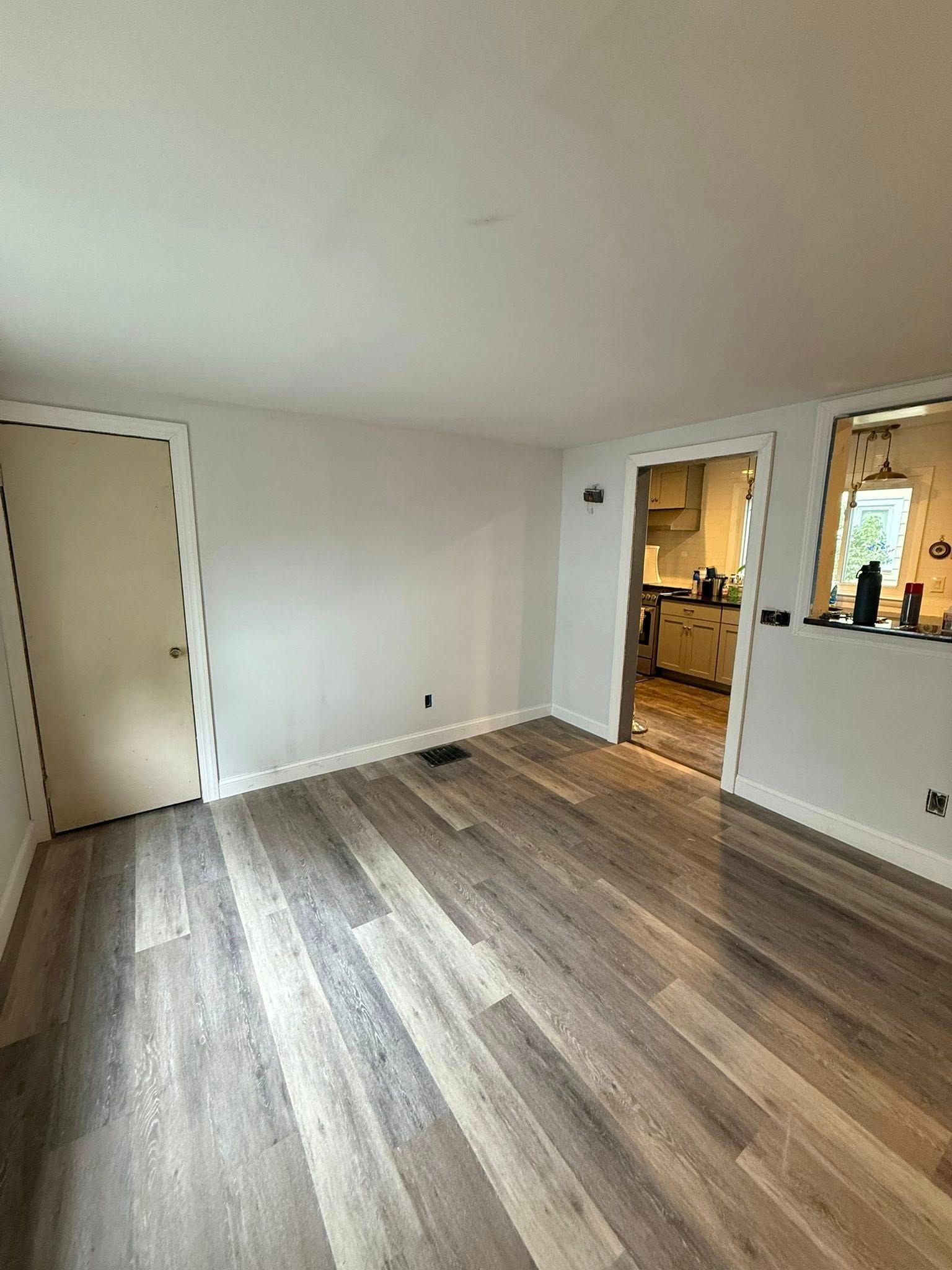 Empty room with wood-look flooring, white walls, and doorways leading to the kitchen and another room.