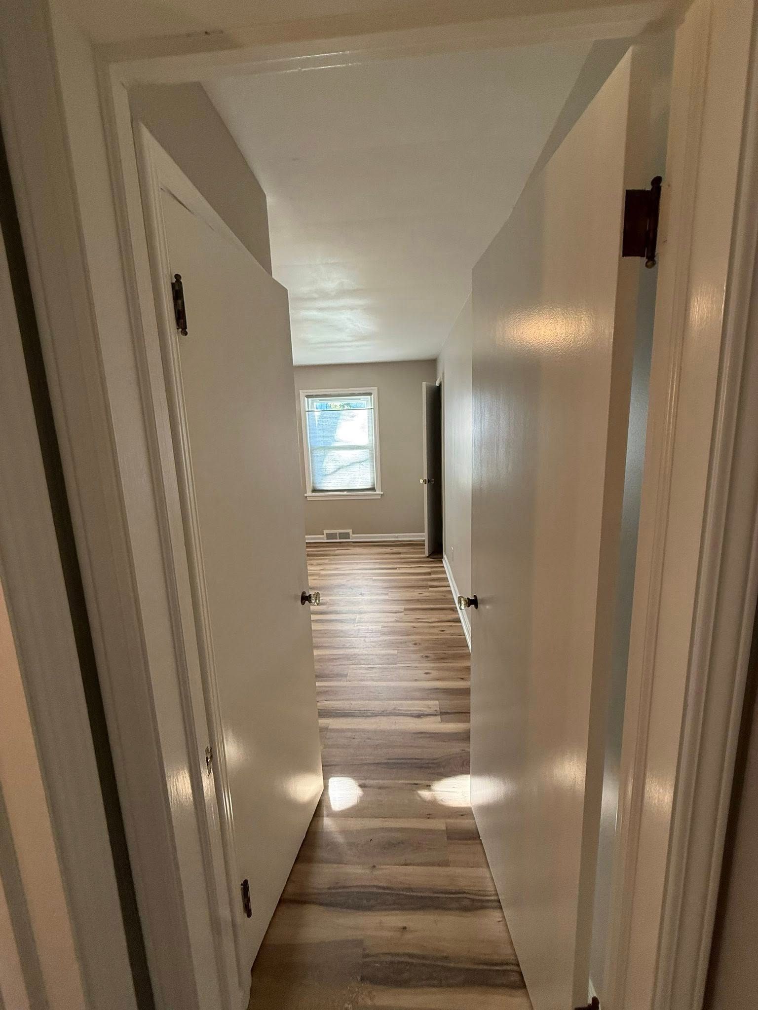 Hallway with light-colored walls and wood-look flooring. Two doors open, revealing a window at the end.