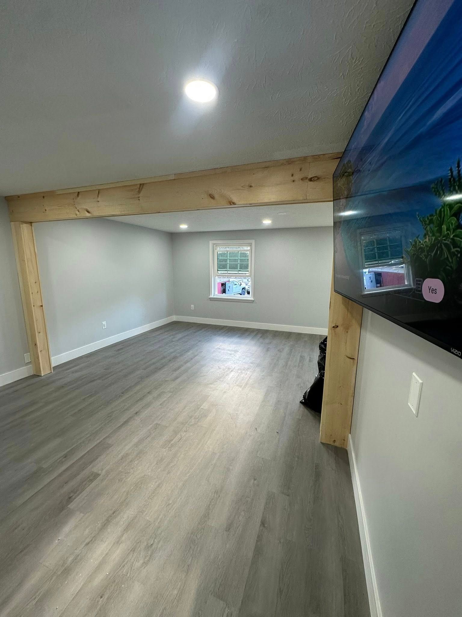 Empty room with gray walls, wood beams, gray flooring, and a window. A TV is mounted on the right wall.