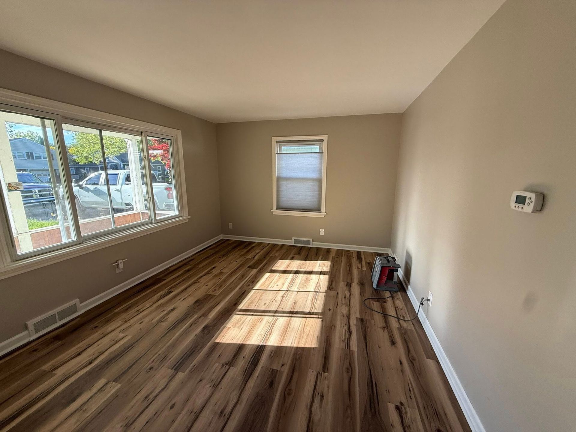 Living room with wood-look flooring, large windows, and neutral walls. Sunlight streams across the floor.