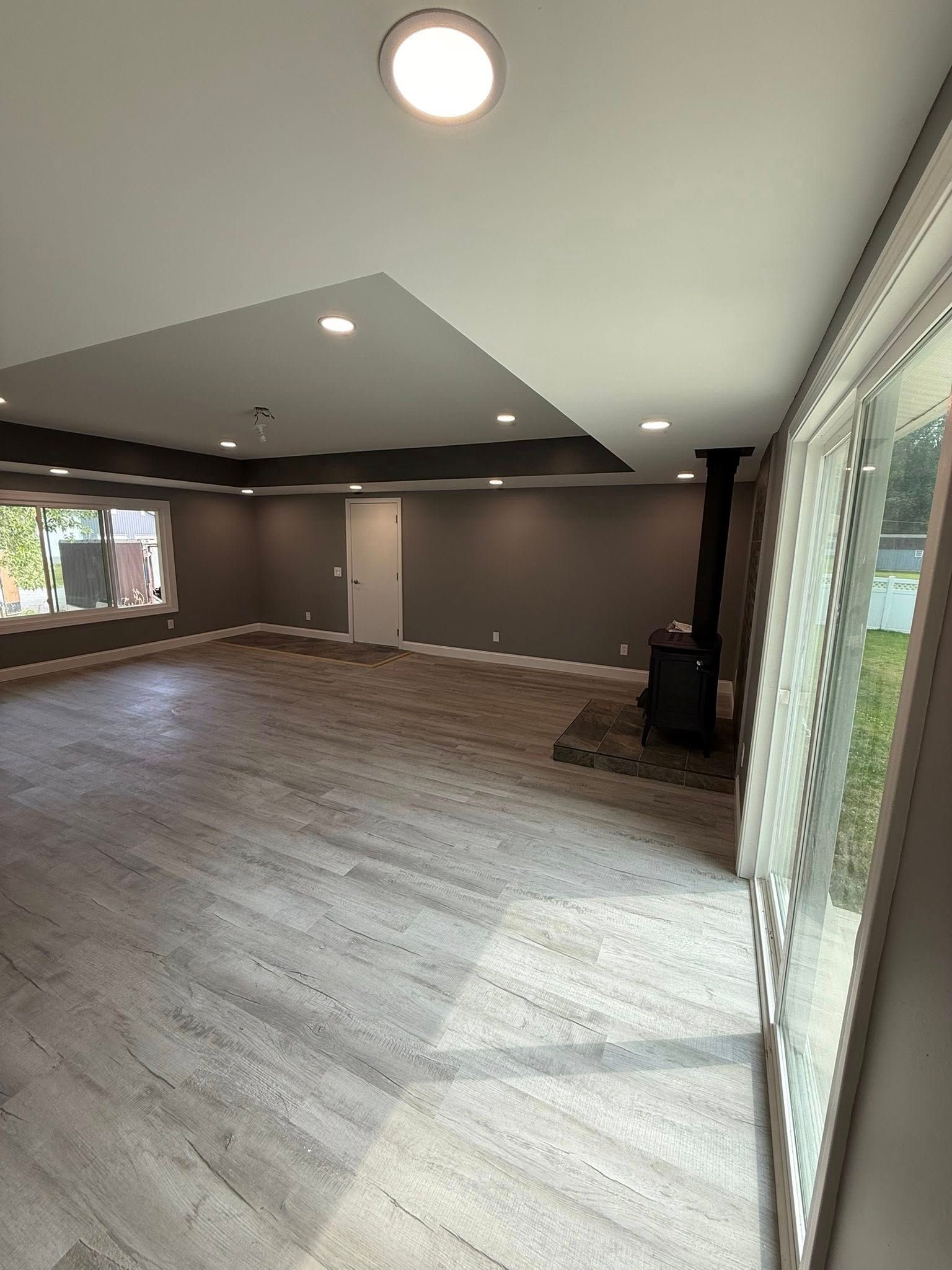 Empty room with wood-look flooring, gray walls and ceiling with recessed lights, wood-burning stove, and sliding glass door.