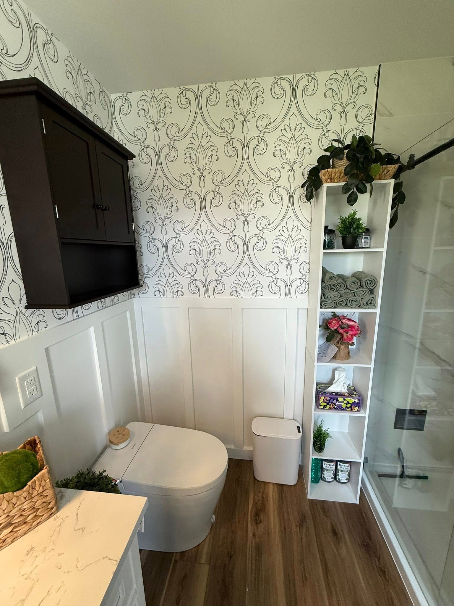 Bathroom with floral wallpaper, white wainscoting, a toilet, storage shelves, and a dark cabinet.
