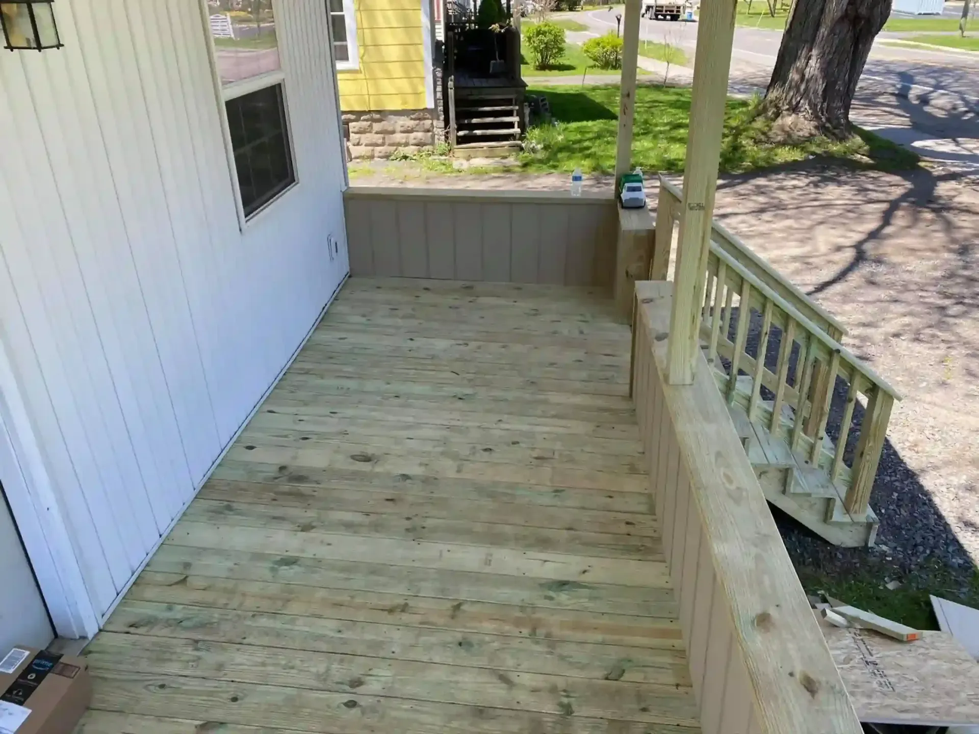 Wooden porch with railing and steps, alongside a white house with a window, and a yellow house in the background.