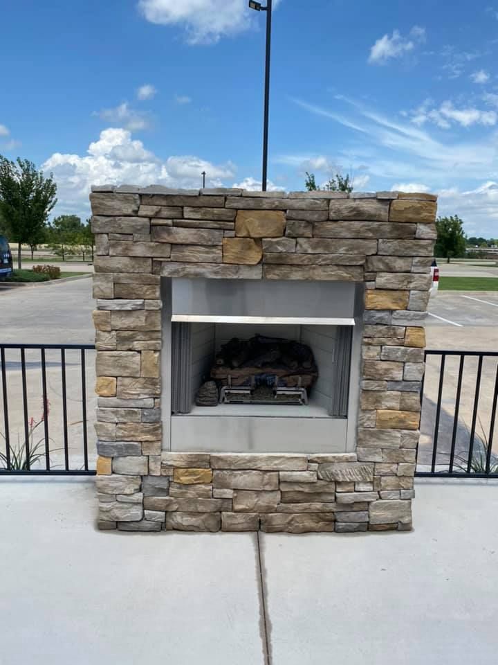 Stone fireplace with a built-in fire feature, outside on a patio, with a metal railing and blue sky. Stone fireplace with a built-in fire feature, outside on a patio, with a metal railing and blue sky.