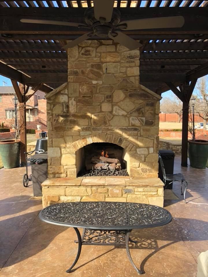 Stone outdoor fireplace with fire, under a pergola, table in foreground.