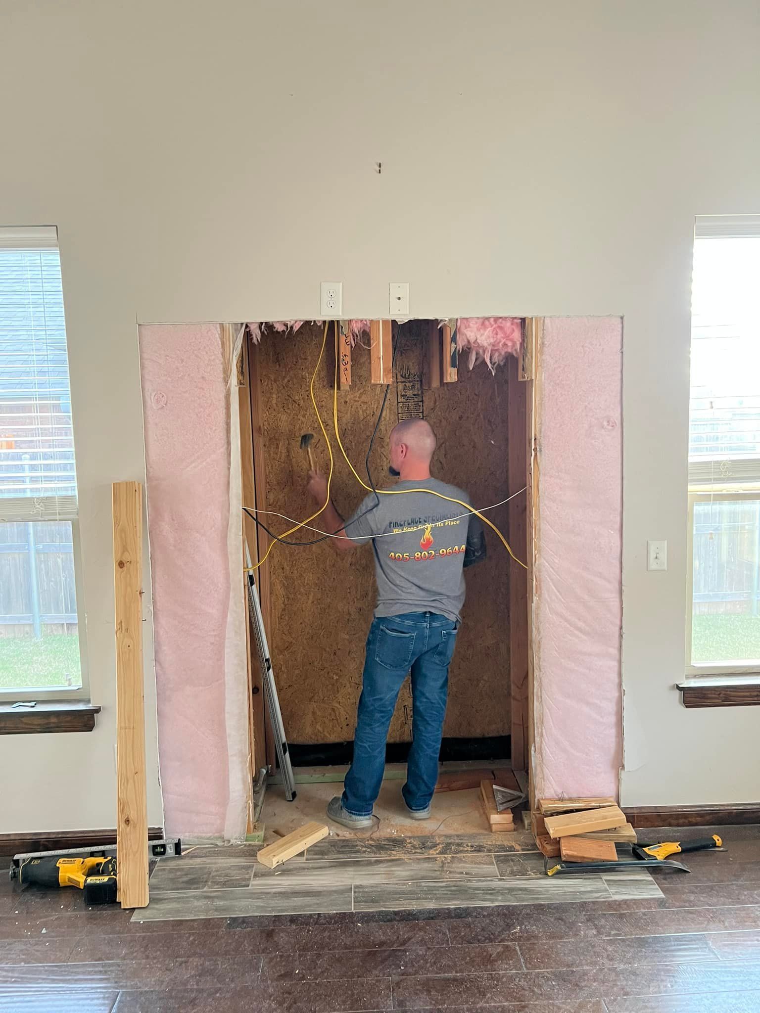 A person working on electrical wiring inside a wall. Pink insulation and construction tools are visible.