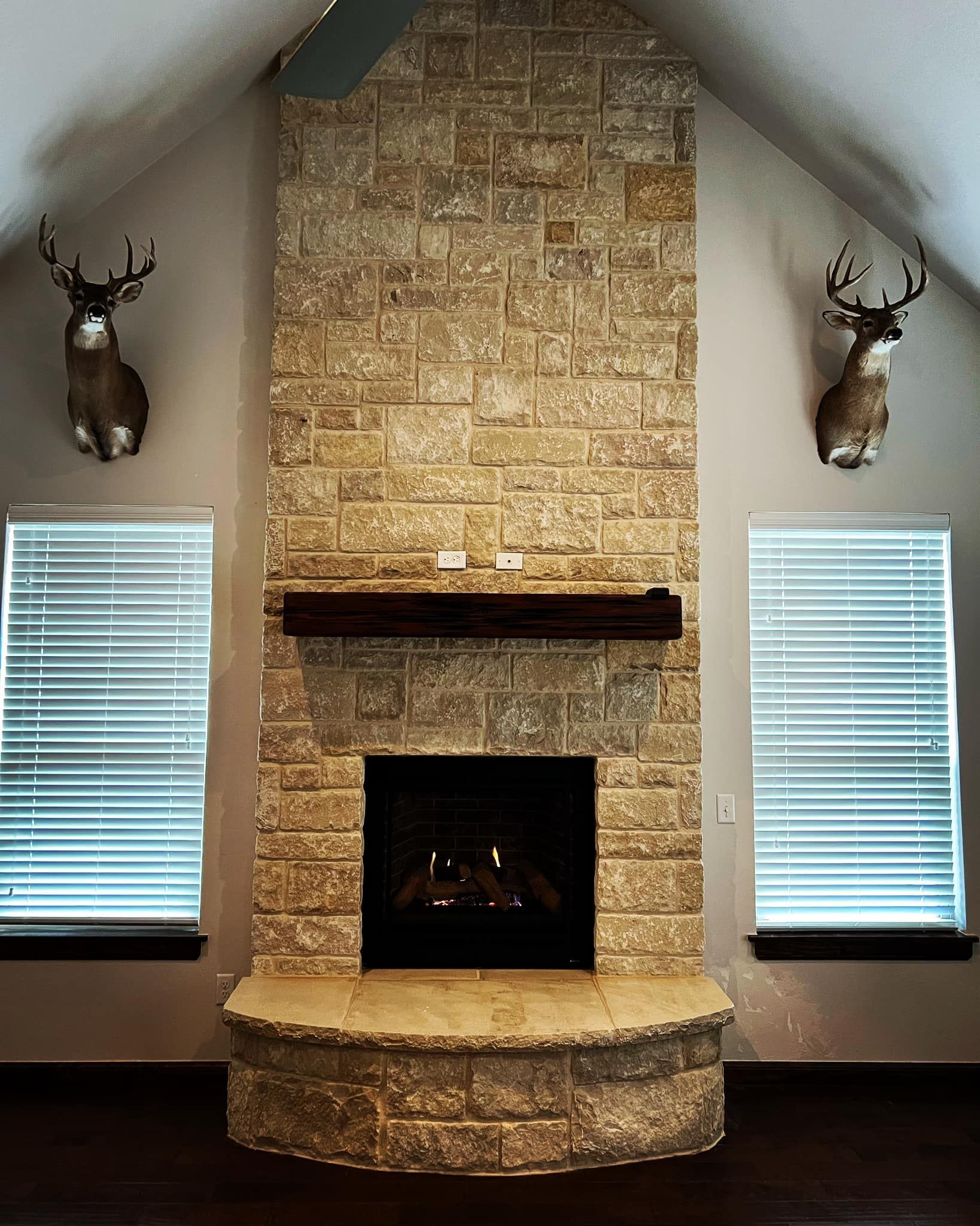 Fireplace with stone facade, dark mantel, flanked by two windows and deer head mounts. Fireplace with stone facade, dark mantel, flanked by two windows and deer head mounts.