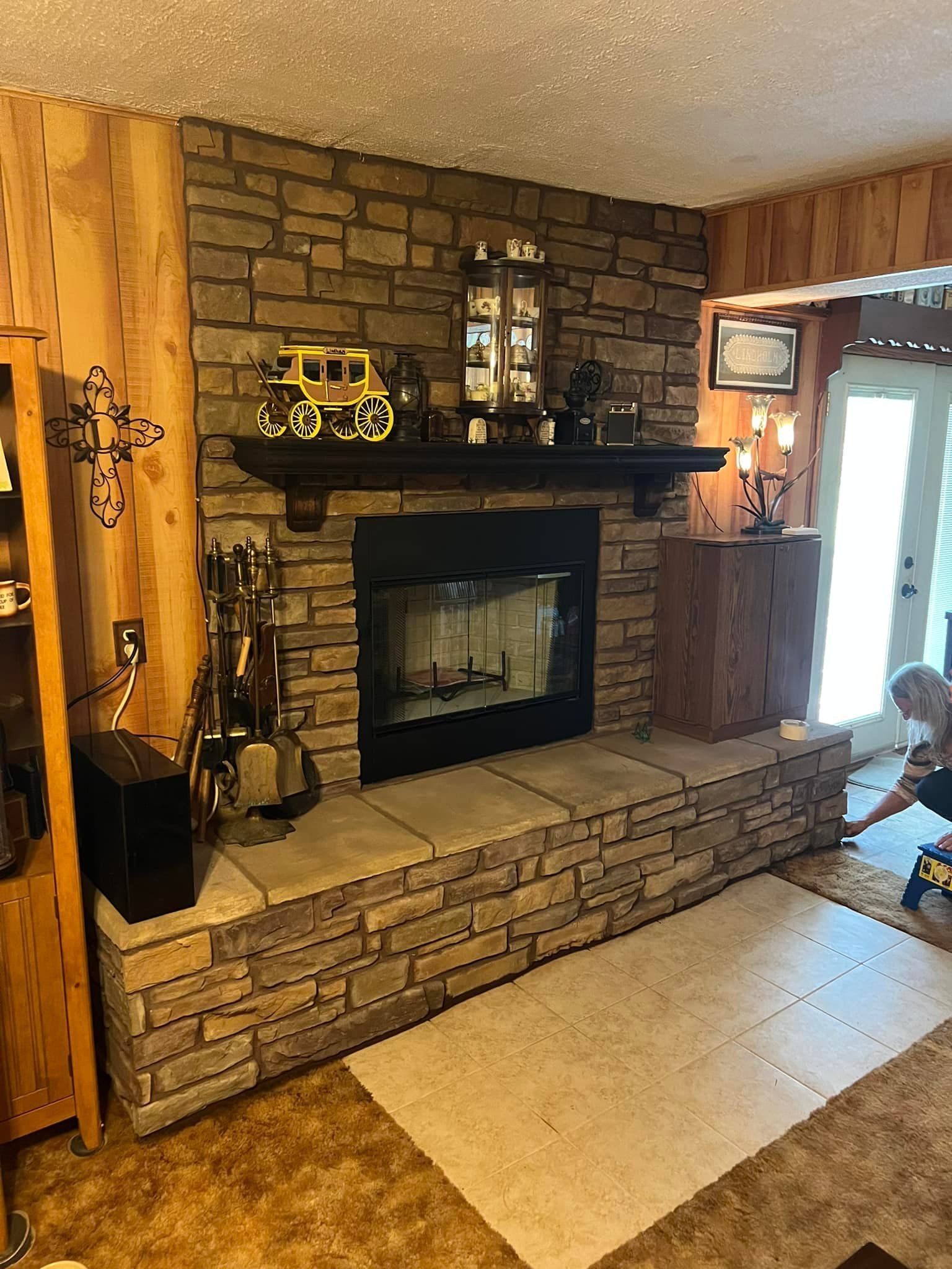 Stone fireplace with black interior and wooden mantel, decorated with figurines. Fireplace hearth extends out, carpet in front. Stone fireplace with black interior and wooden mantel, decorated with figurines. Fireplace hearth extends out, carpet in front.