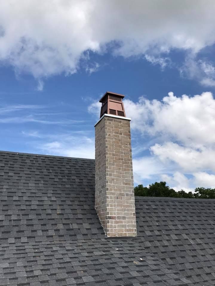 Brick chimney on a dark shingled roof against a blue sky with clouds.