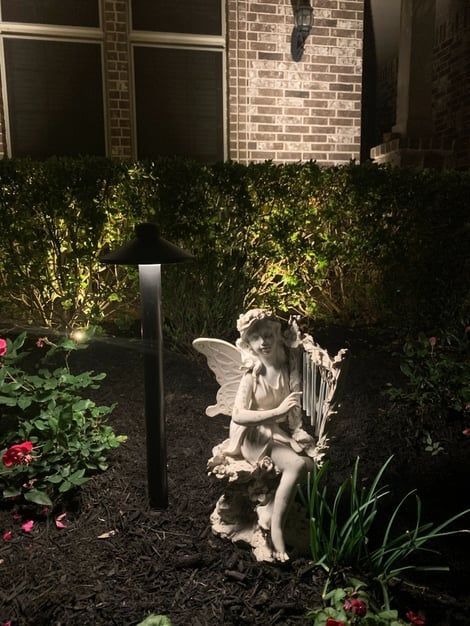 A garden statue of an angel with wings sits next to a path light and greenery at night.