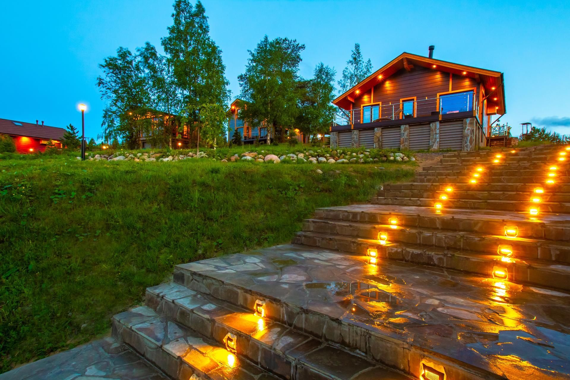 Stone steps illuminated with lights lead up to a wood cabin at dusk.