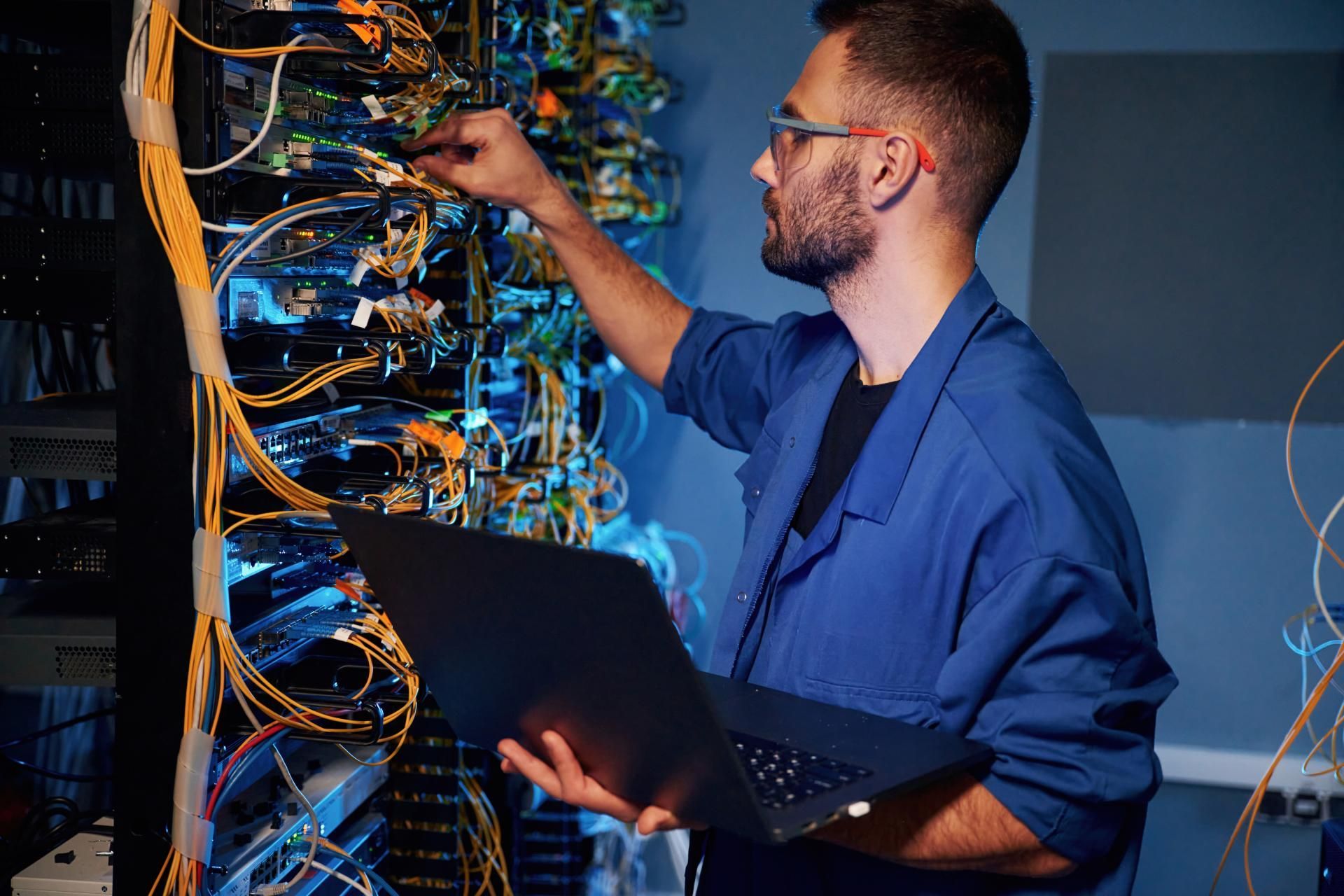 Technician in blue coveralls using laptop while working on server hardware in a server room.