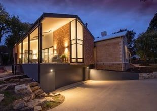 Modern home at dusk with glass walls, brick facade, and lit driveway.