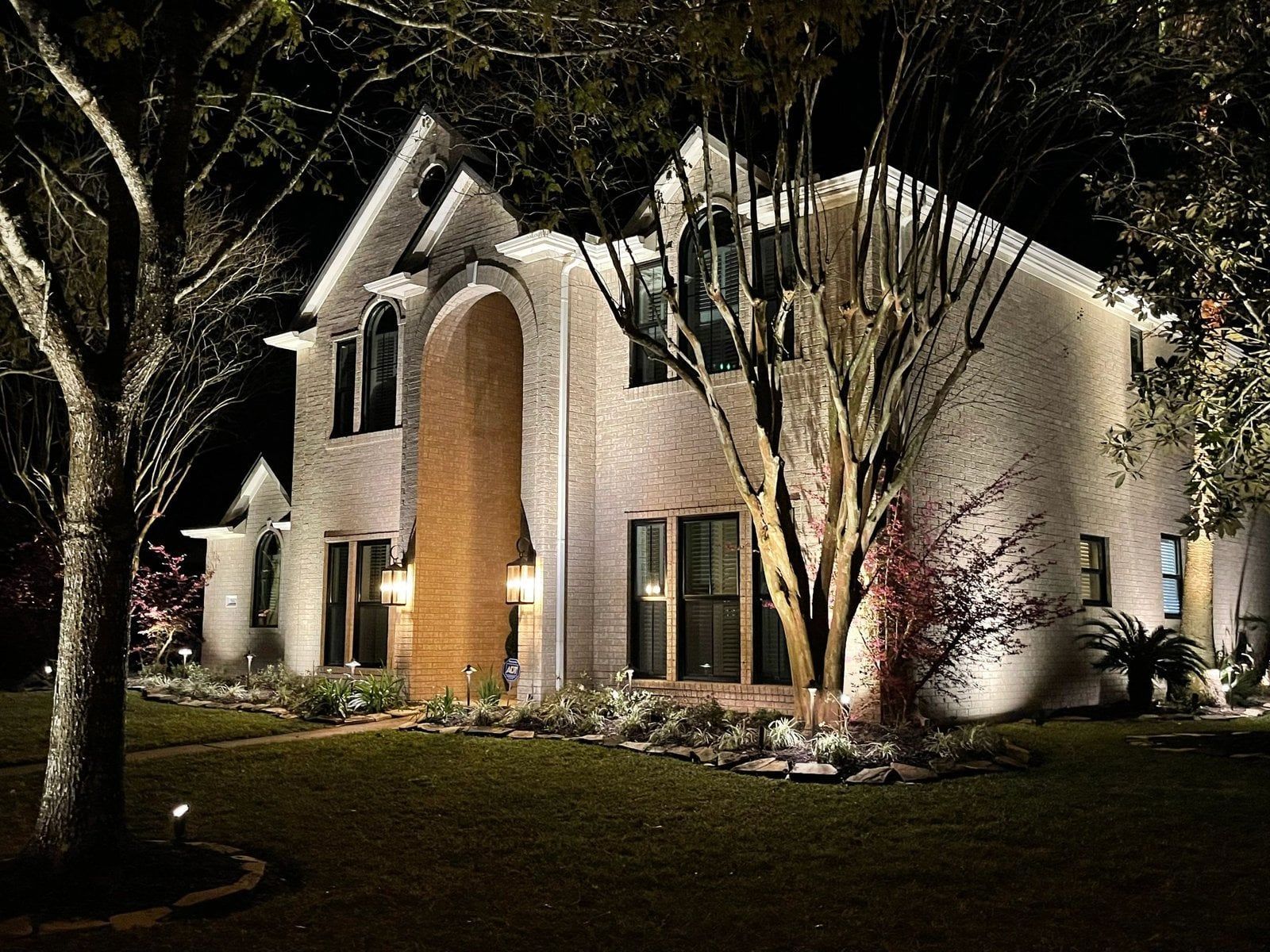 Night view of a two-story brick house illuminated by outdoor lighting. Trees and lawn in foreground.