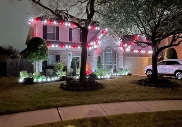 House decorated with red and white Christmas lights, trees, and a car in the driveway at night.