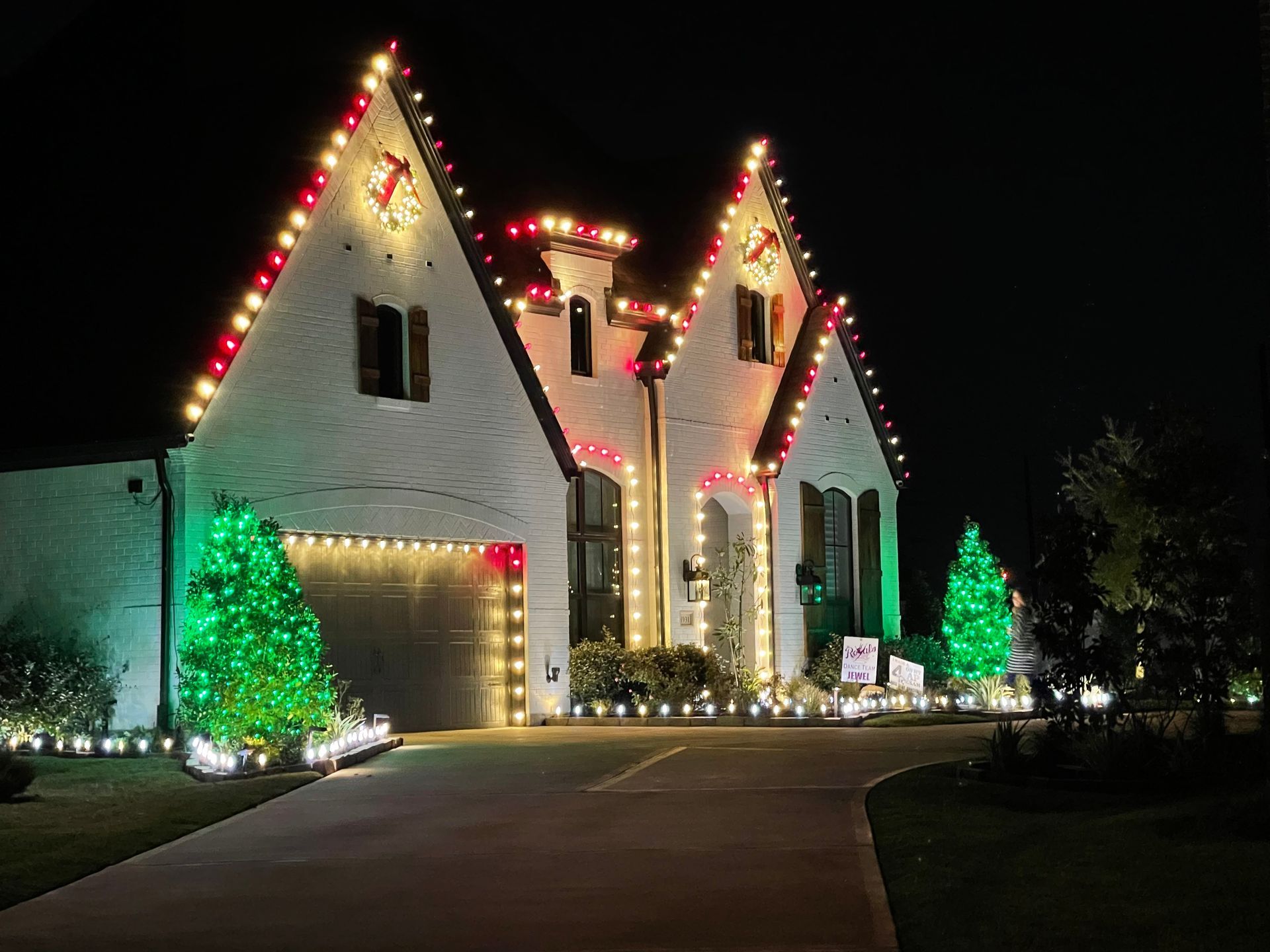 House decorated with Christmas lights at night; red and white lights on the roof, green lights on bushes.