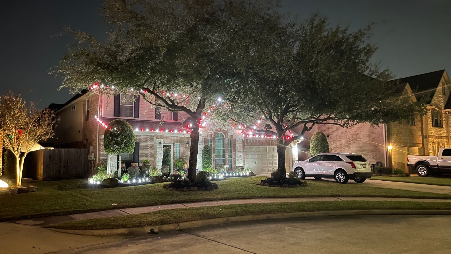 House decorated with red Christmas lights, illuminated trees, and white SUV parked in the driveway at night.