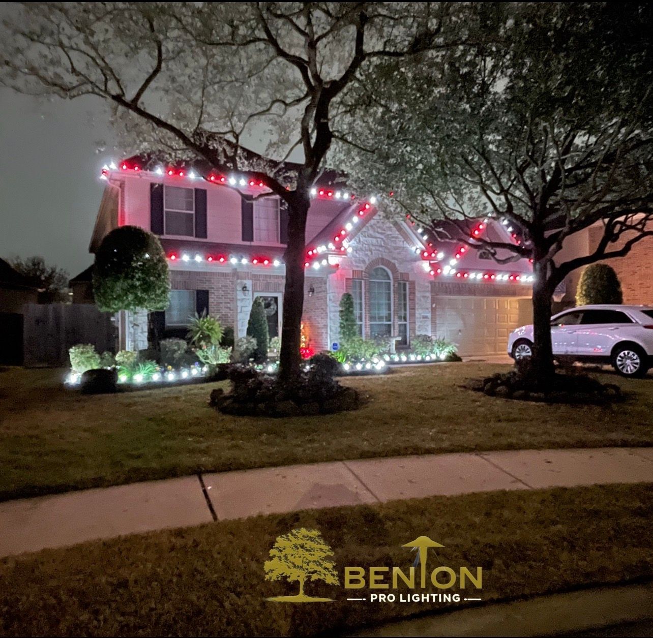 A two-story house with red and white Christmas lights and illuminated landscaping at night.