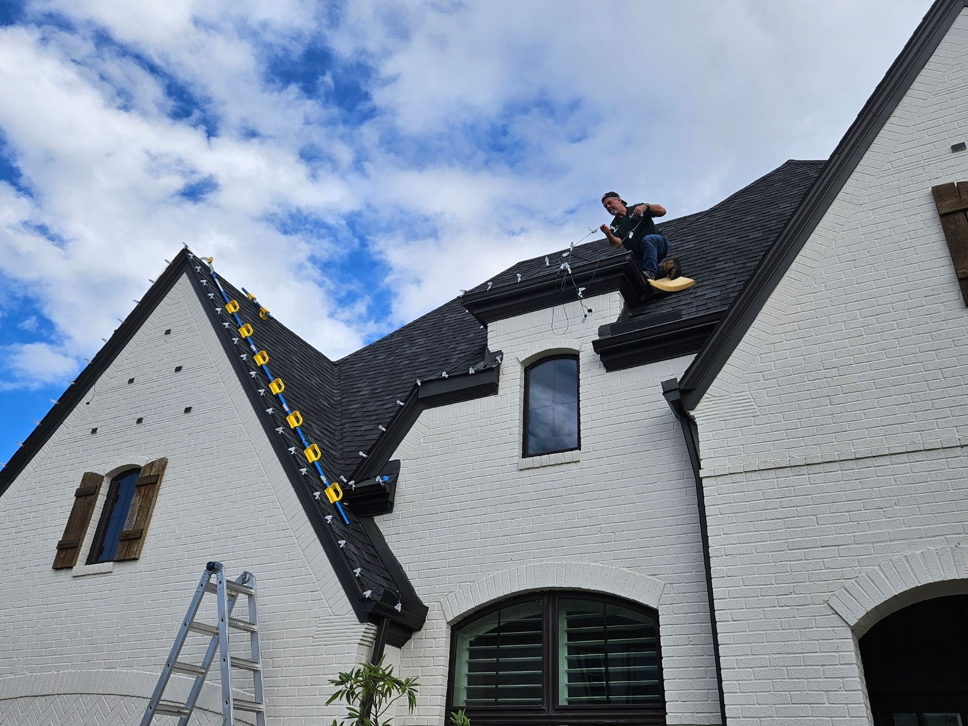 Person on a roof installing holiday lights on a white brick house under a cloudy sky.