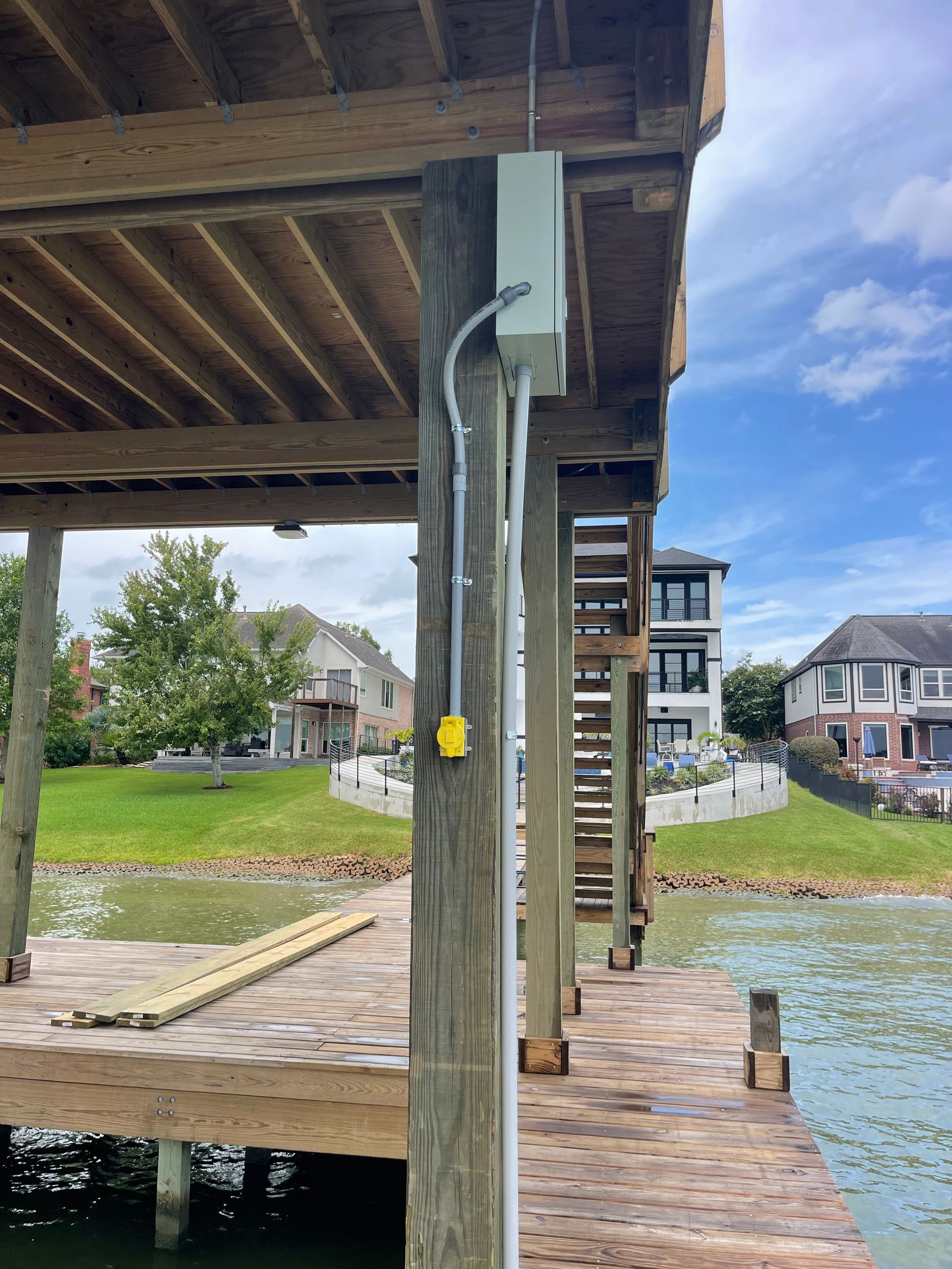 Wooden pier with electrical conduit and outlet on a support post, waterfront with houses in the background.