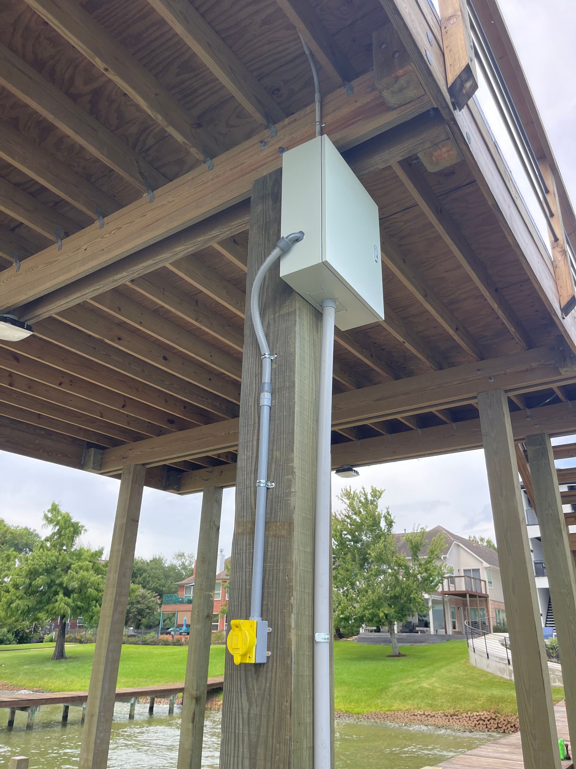 Electrical box and conduit on a wooden post under a dock.  Outlet box on the post, green grass, water, and homes in the background.