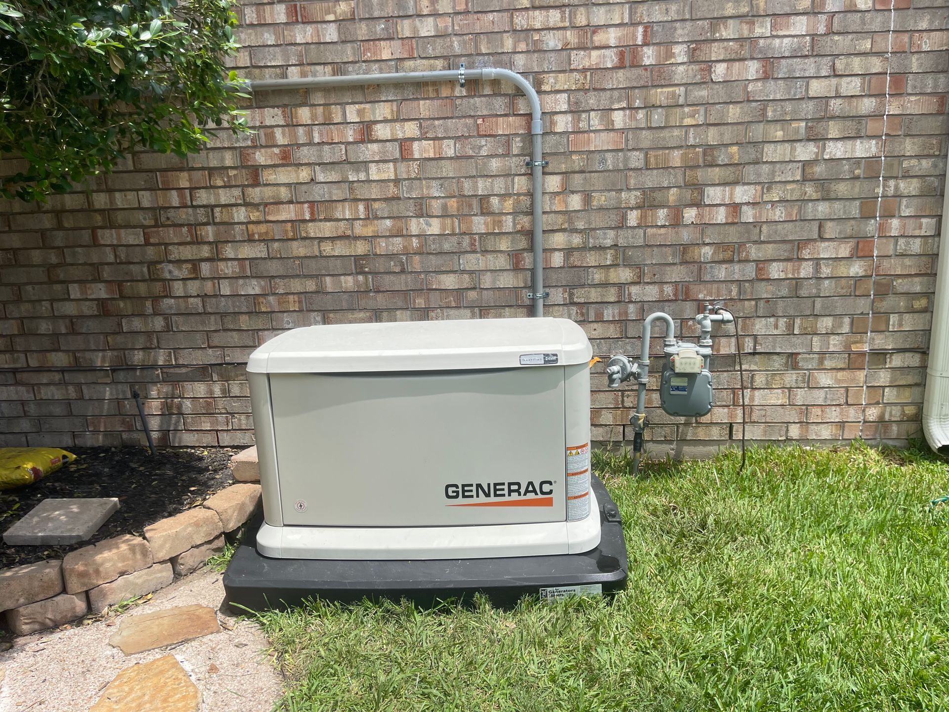 Generac generator against a brick wall, next to a gas meter, on a black pad, and on grass.